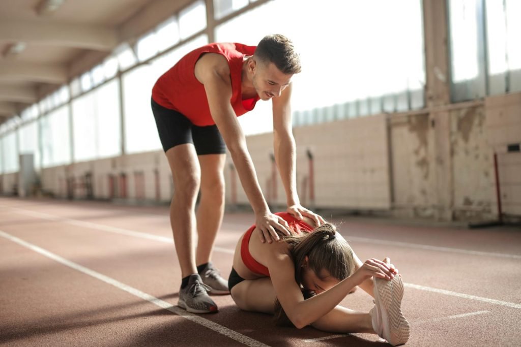 Miriampdmoda Coaching Marbella - Professional male sportsman helping woman touching knees with head while female sitting on running track and doing stretching exercise